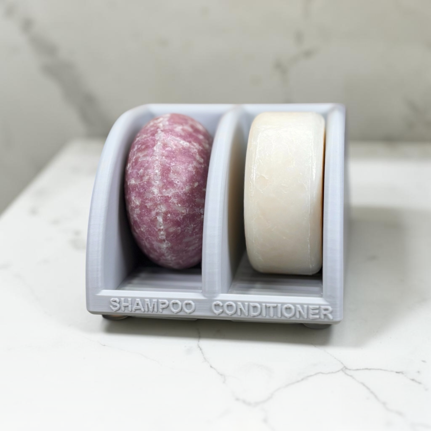 Two soap bars, one pink and one white, in a light gray soap dish on a marble surface.