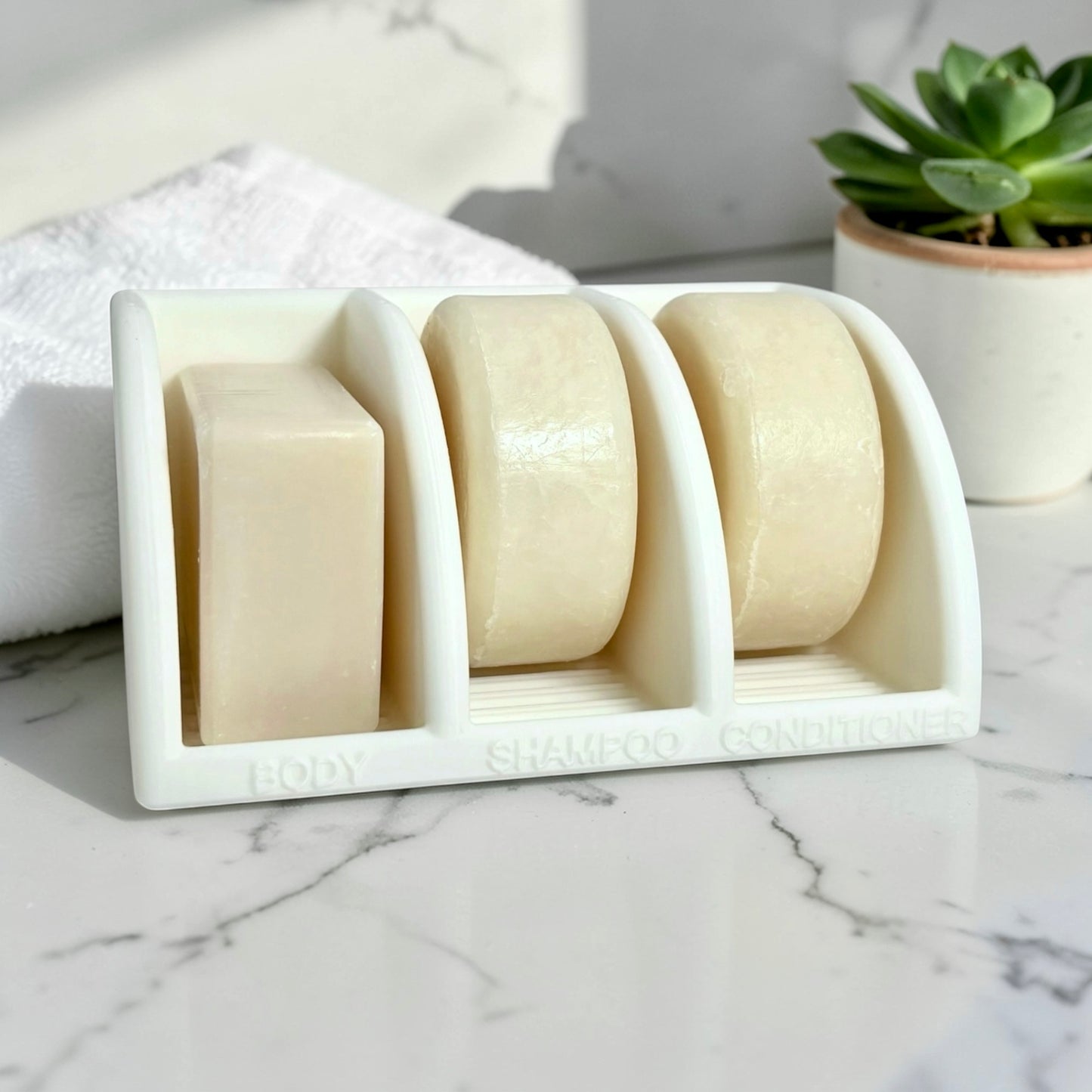 Three bars of soap in a white soap dish on a marble surface with a plant in the background.