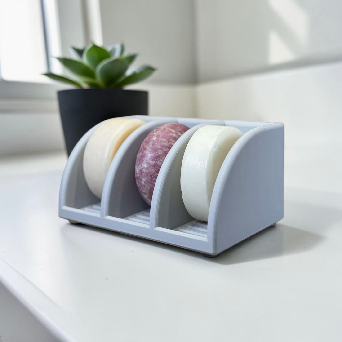 Three soap bars in a light gray holder on a light surface with a plant in the background