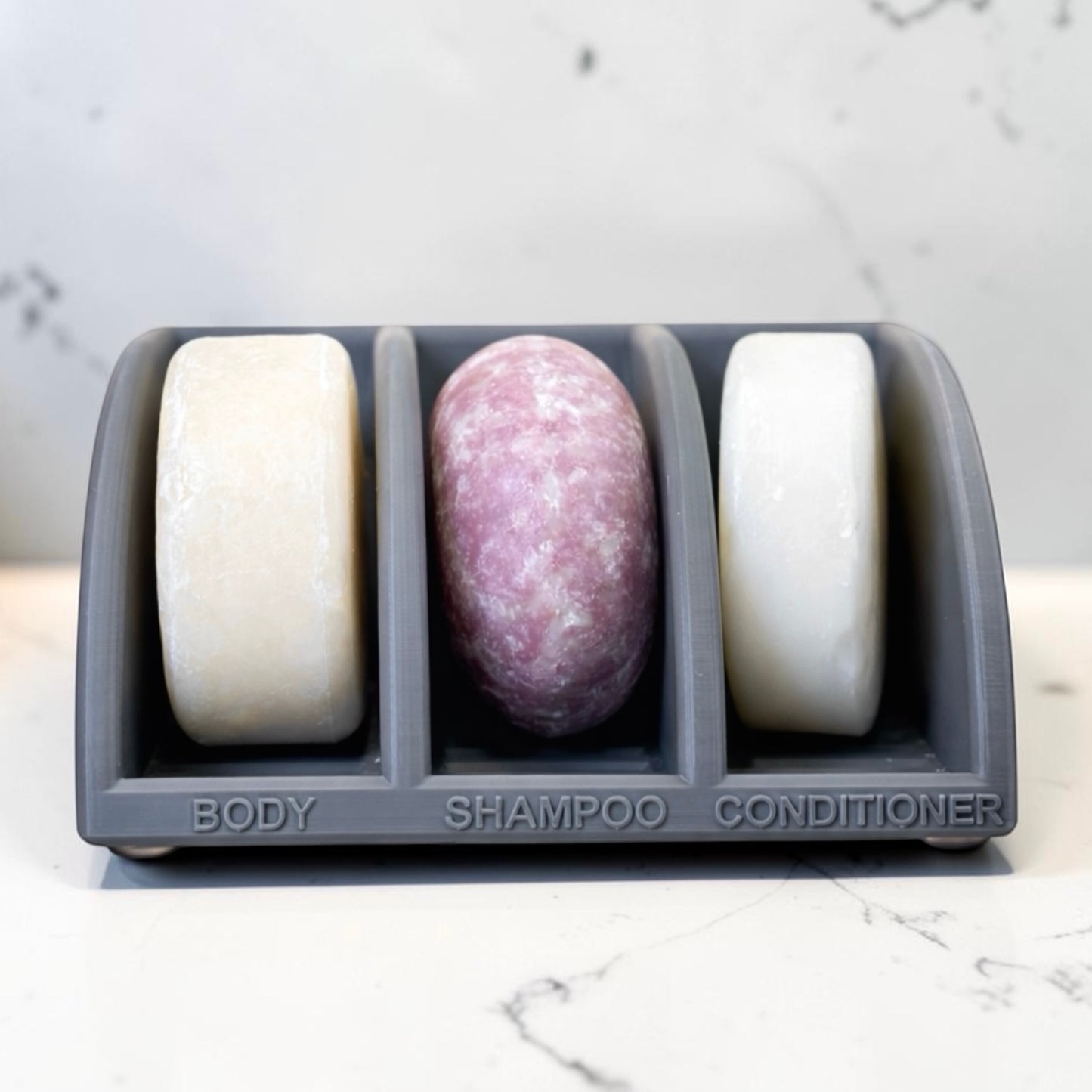 Three soap bars in a gray holder labeled 'Body', 'Shampoo', and 'Conditioner' on a marble surface.