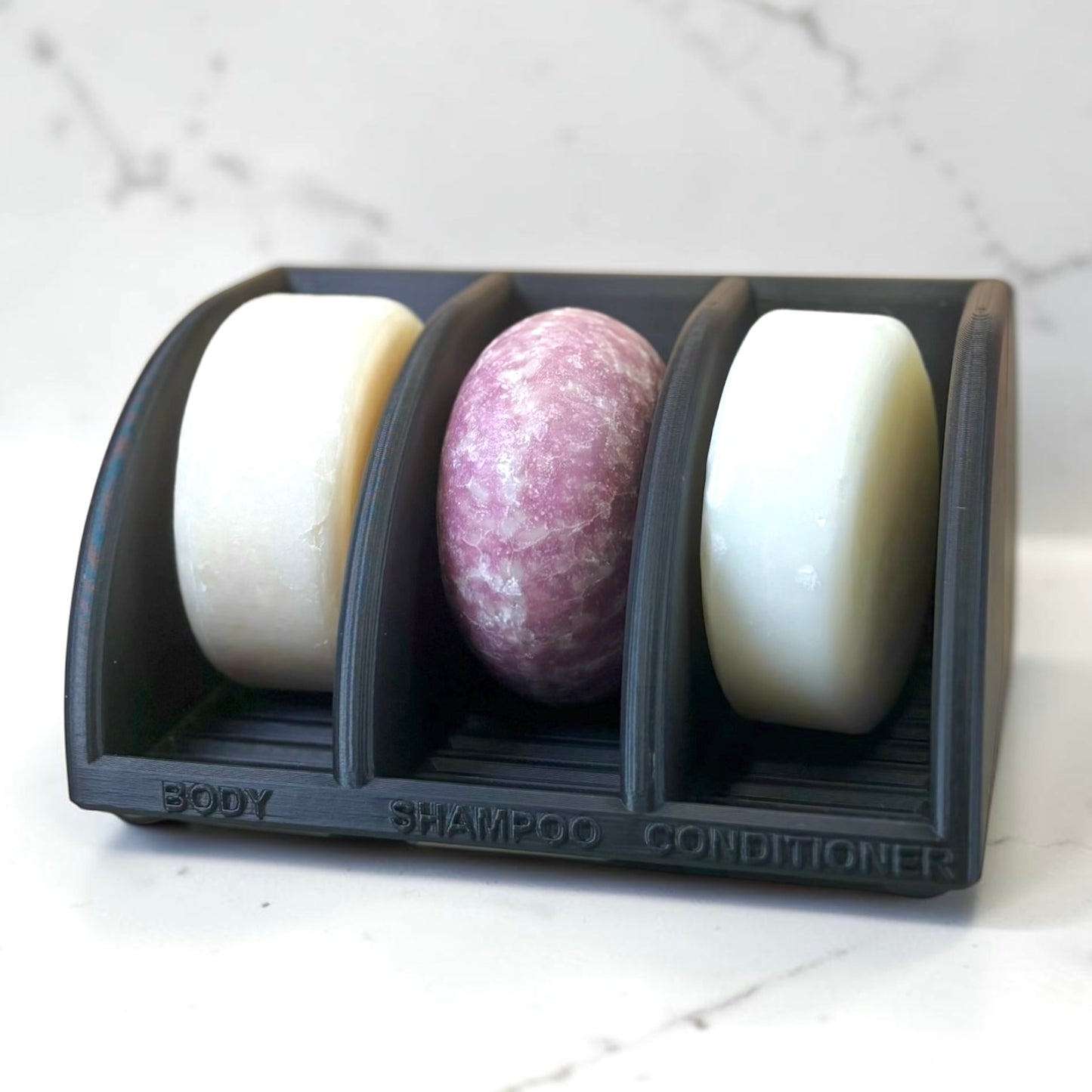 Three bars of soap in a black holder labeled 'Body', 'Shampoo', and 'Conditioner' on a white marble background.