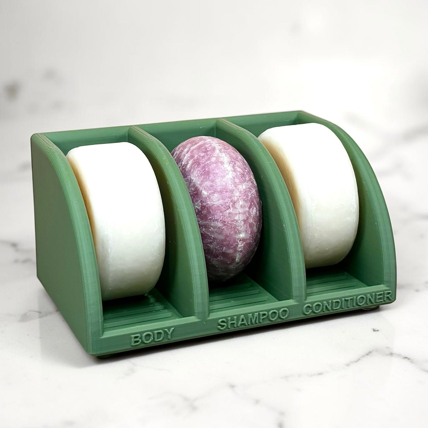 olive green soap holder with three compartments labeled 'Body', 'Shampoo', and 'Conditioner' on a marble surface.