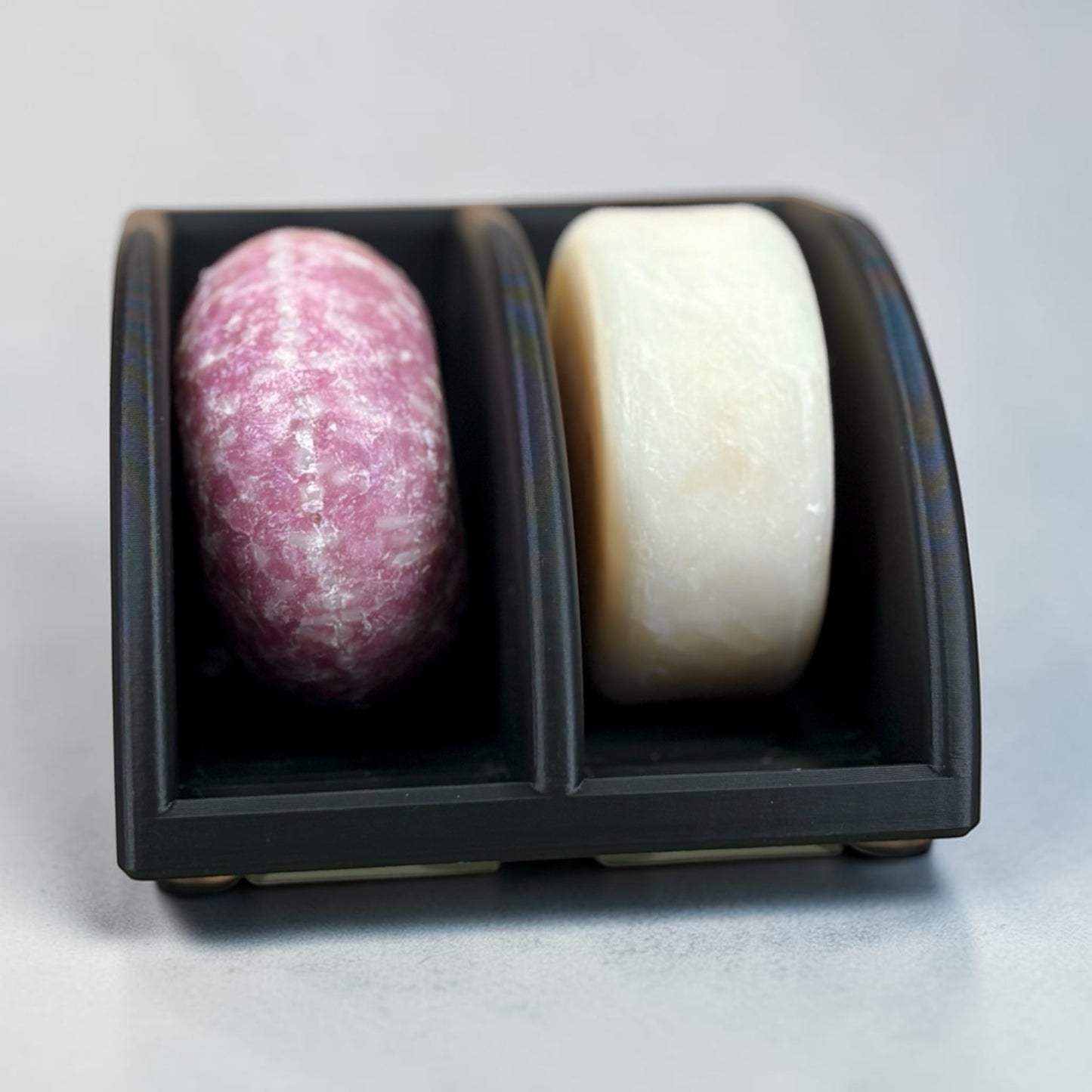 Two soap bars, one pink and one white, in a black soap dish on a light gray background.