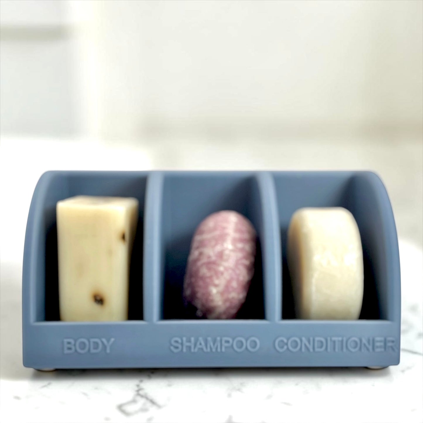 Three bars of soap in a compartmentalized holder labeled 'Body', 'Shampoo', and 'Conditioner' on a marble surface.