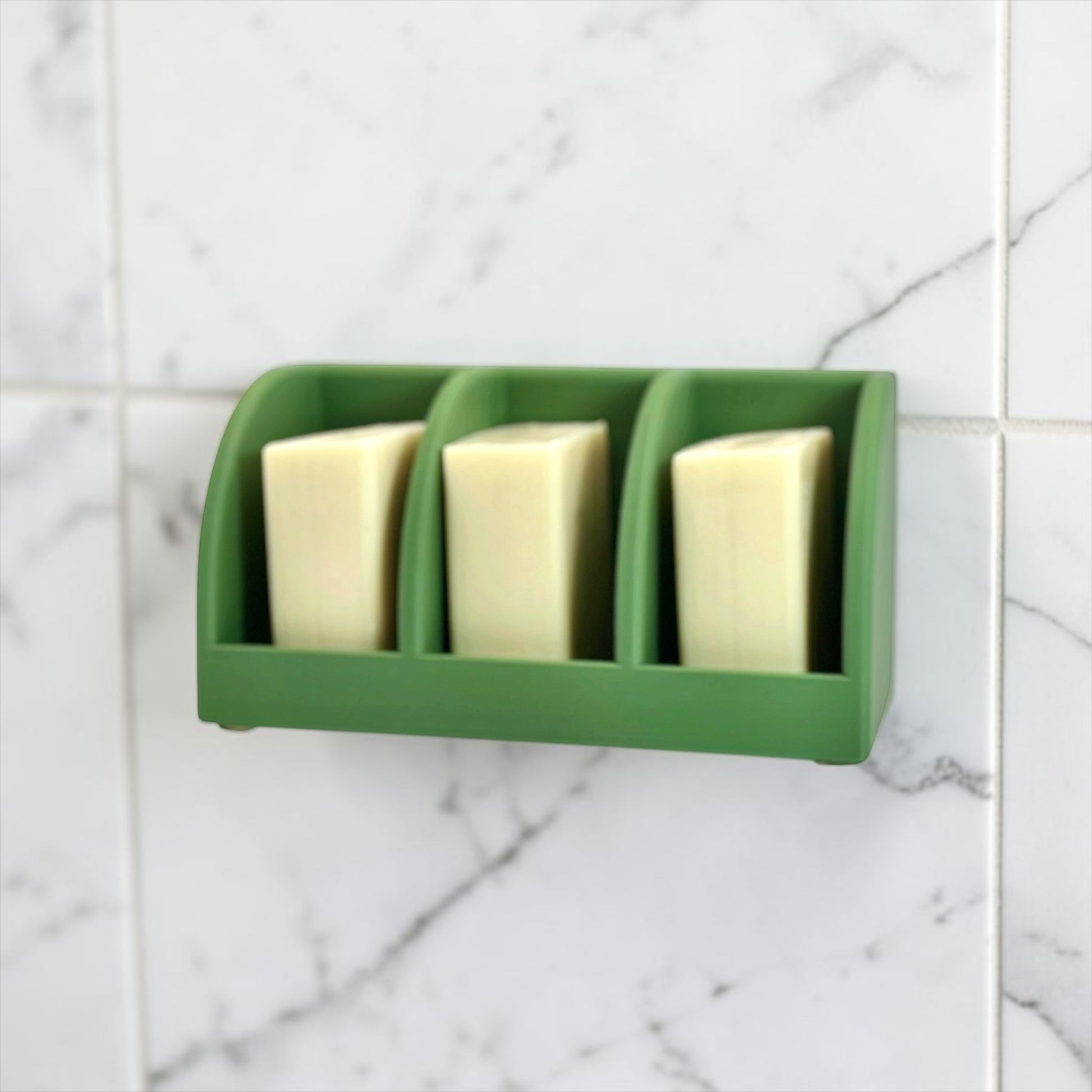 Green soap holder with three bars of soap on a tiled bathroom wall.