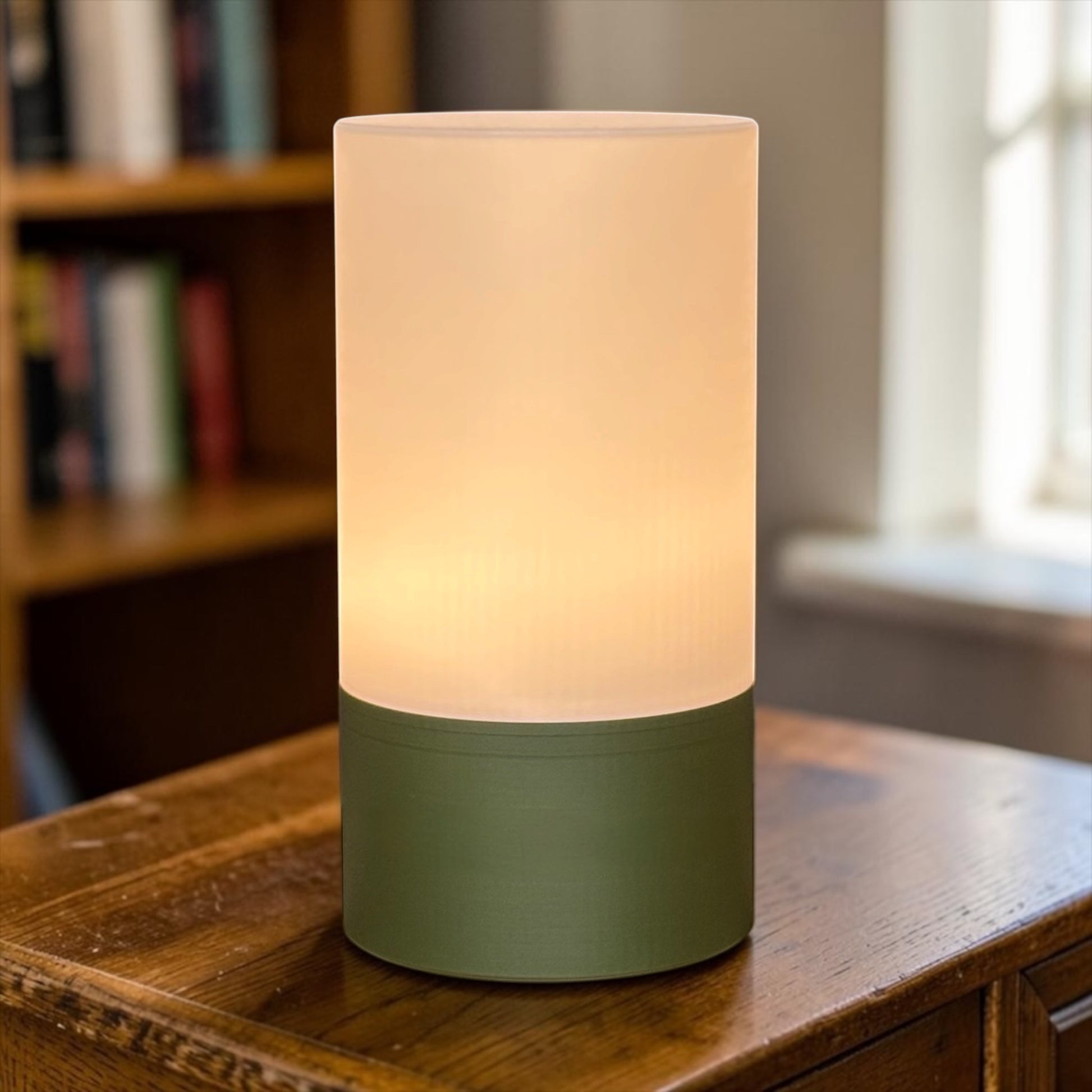 Cylindrical table lamp with a green base on a wooden surface, blurred bookshelf and window in the background