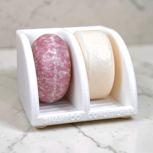 Two bars of soap, one pink and one white, in a white soap dish on a marble surface.