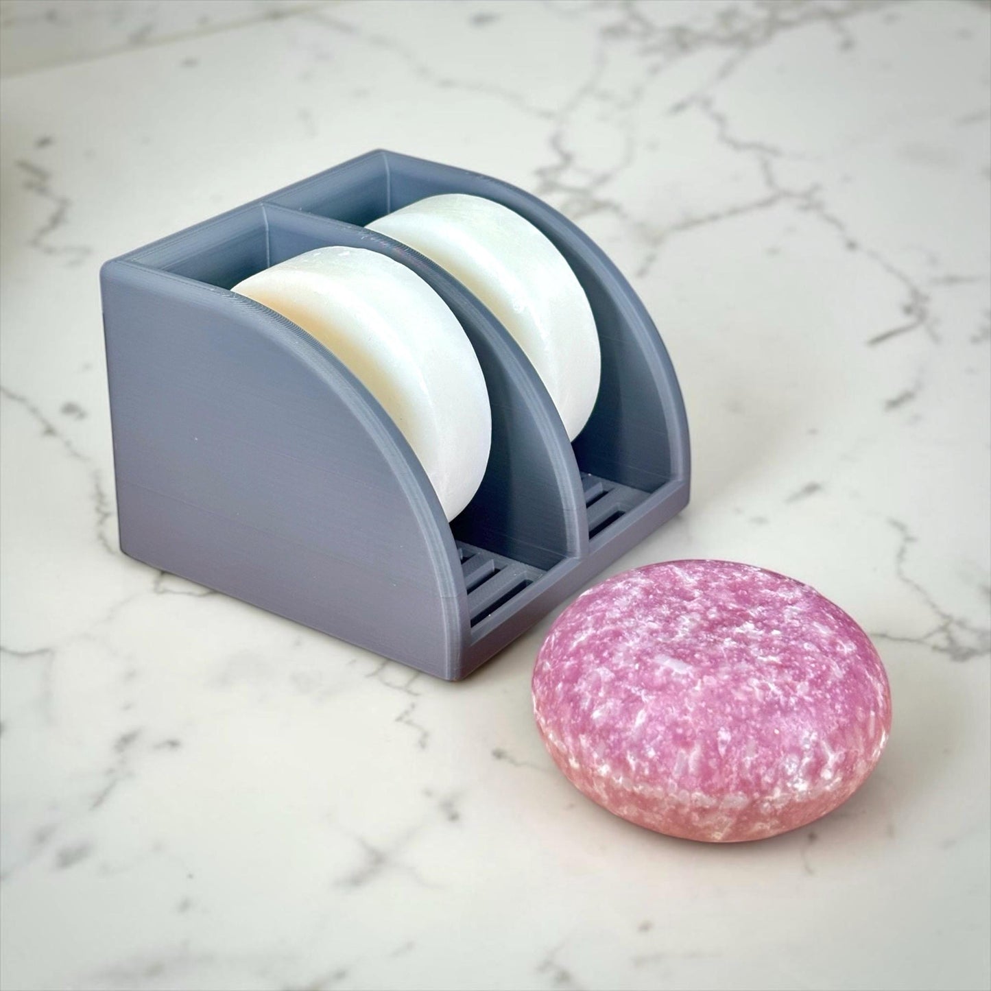 Dark Gray soap holder with white bars of soap and a pink speckled bar on a marble surface