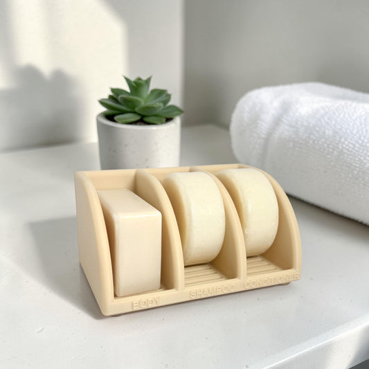 soap holder with three bars on a white surface with a plant and towel in the background.