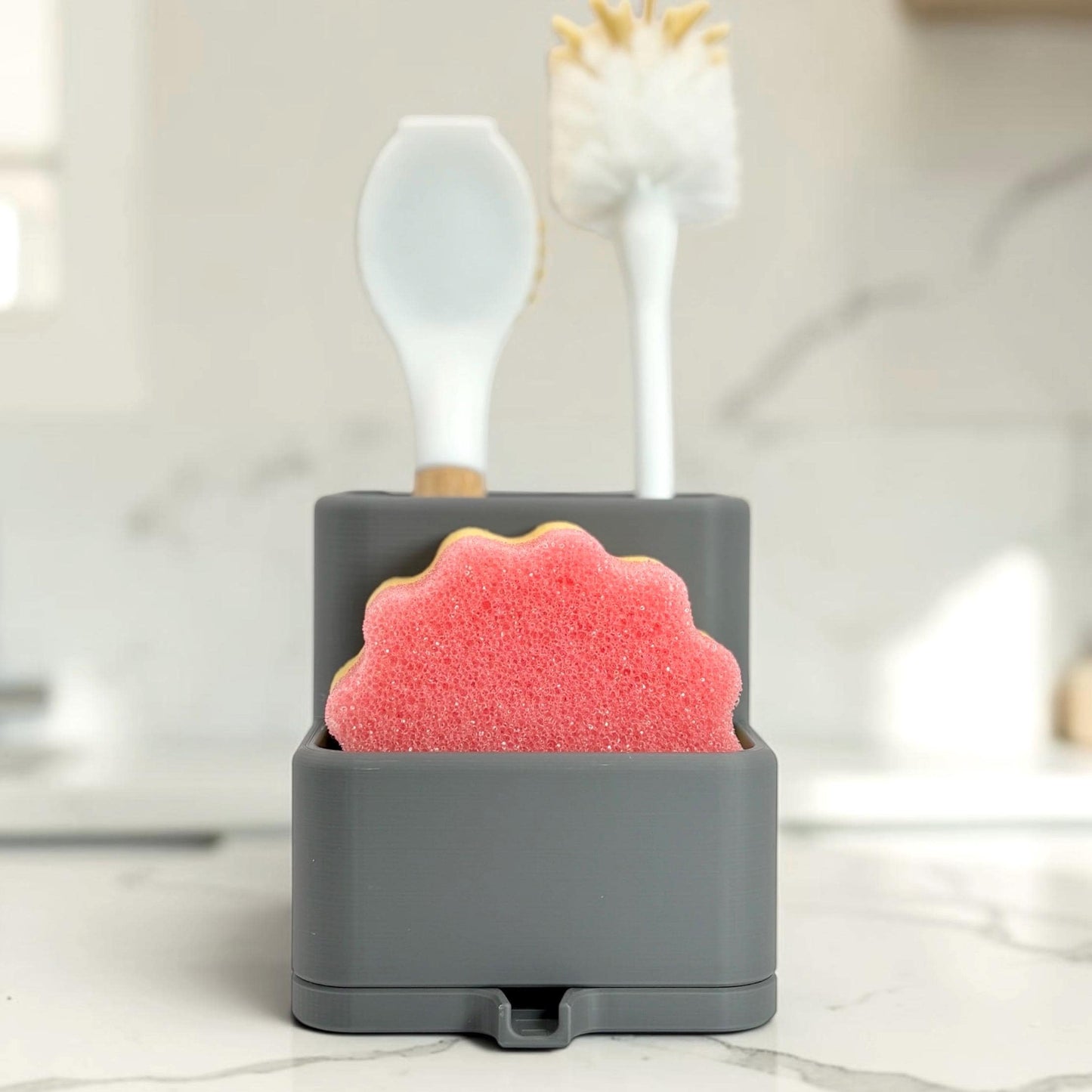 Gray utensil holder with a pink sponge and white spoon on a marble surface.