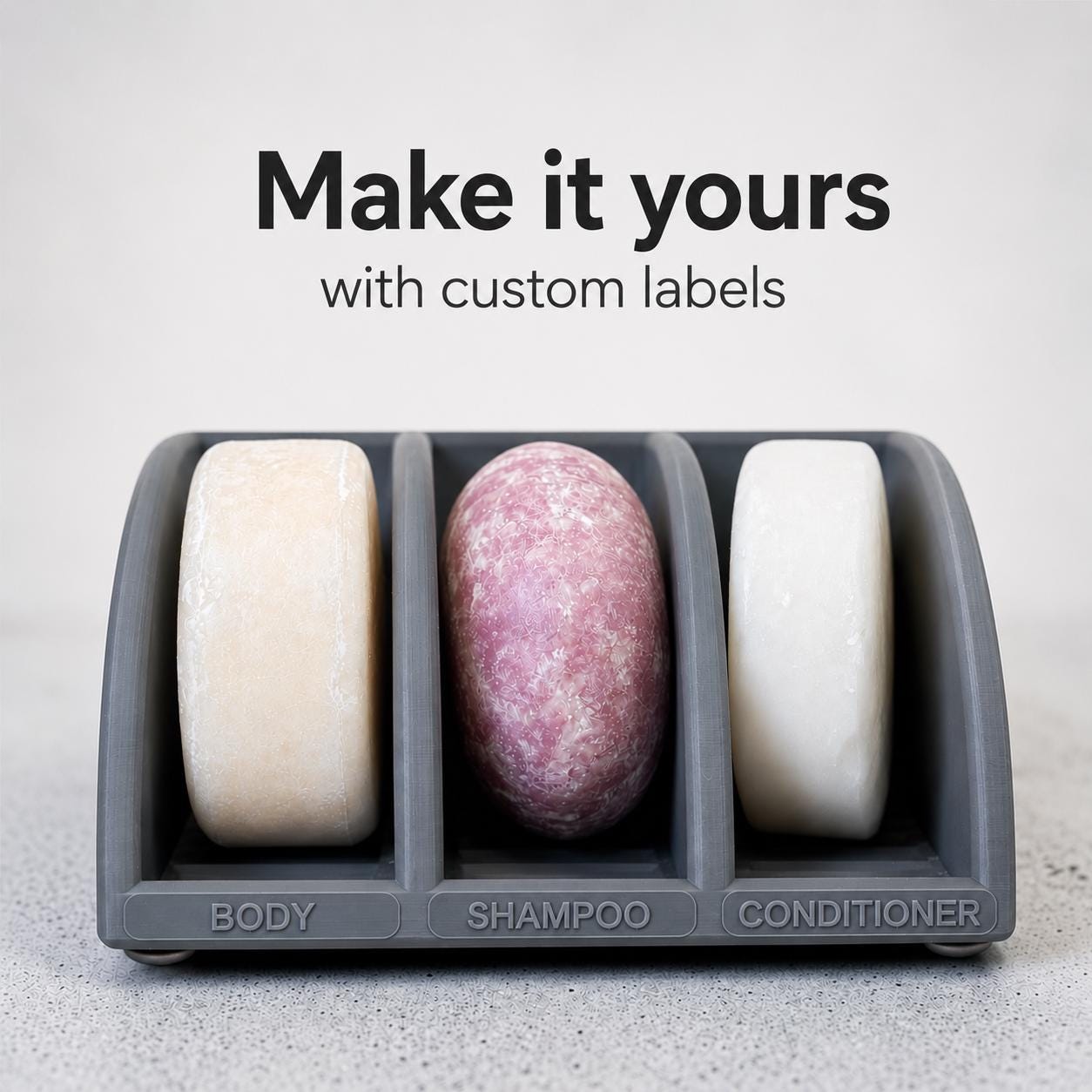 Three soap bars in a holder labeled 'Body', 'Shampoo', and 'Conditioner' with text 'Make it yours with custom labels' on a light background.
