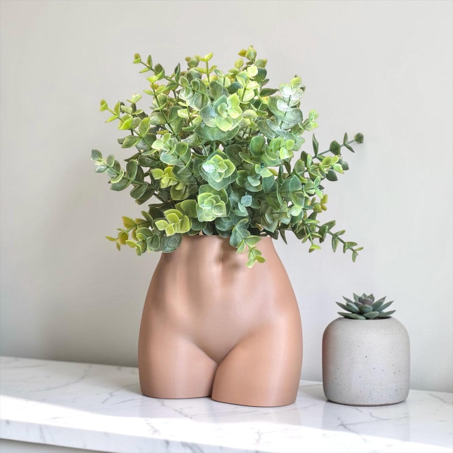 Vase shaped like a female torso with green plant and small succulent on a marble surface.